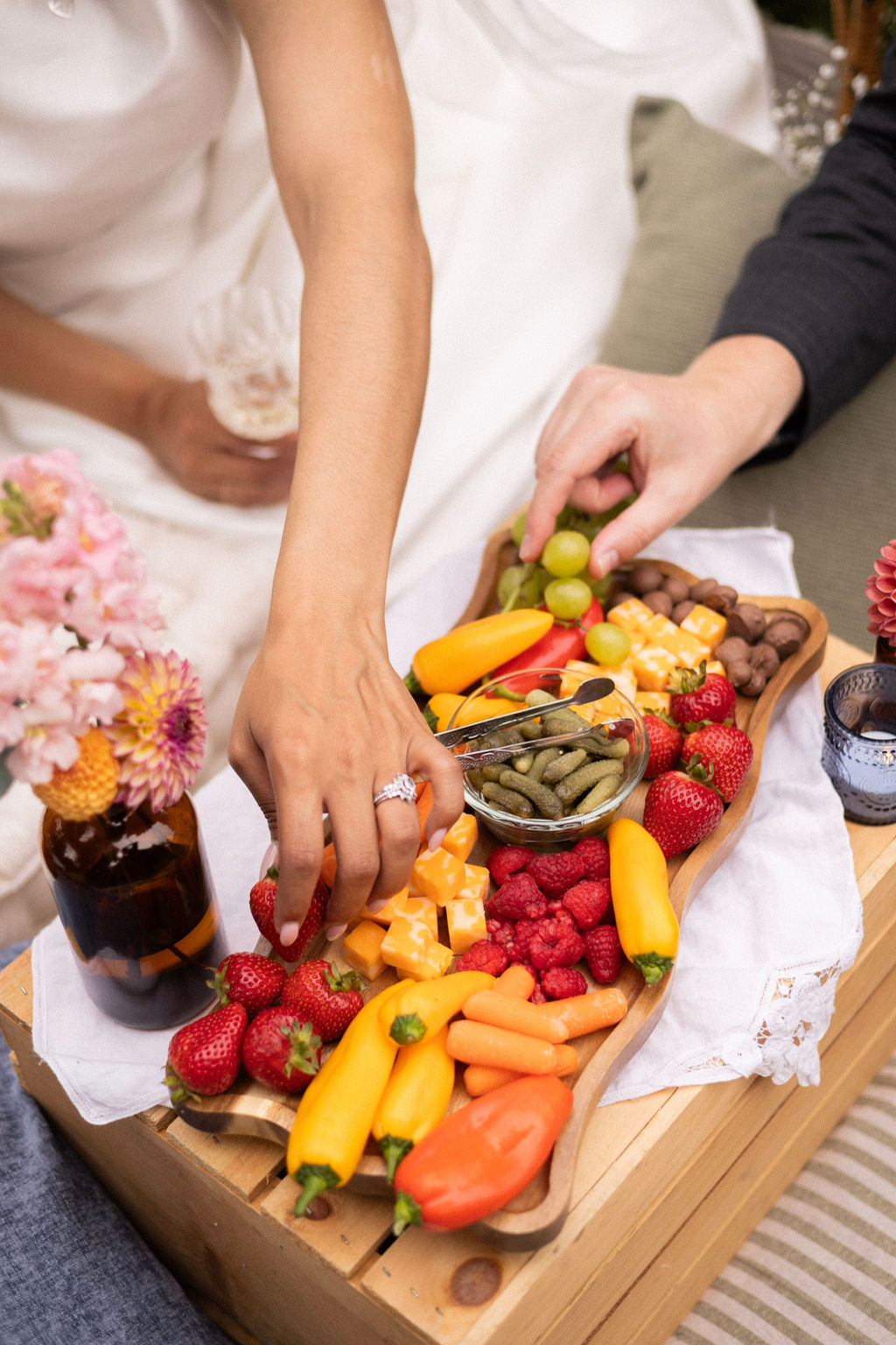 Two hands reaching for a grazing board of strawberries, peppers, cheese and grapes at a blanket picnic