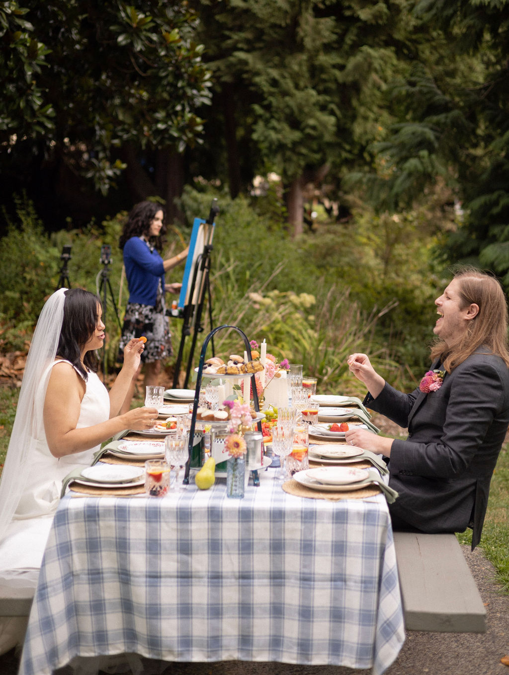 Newlyweds laughing at the picnic table while the live event painter, an add-on, works behind them