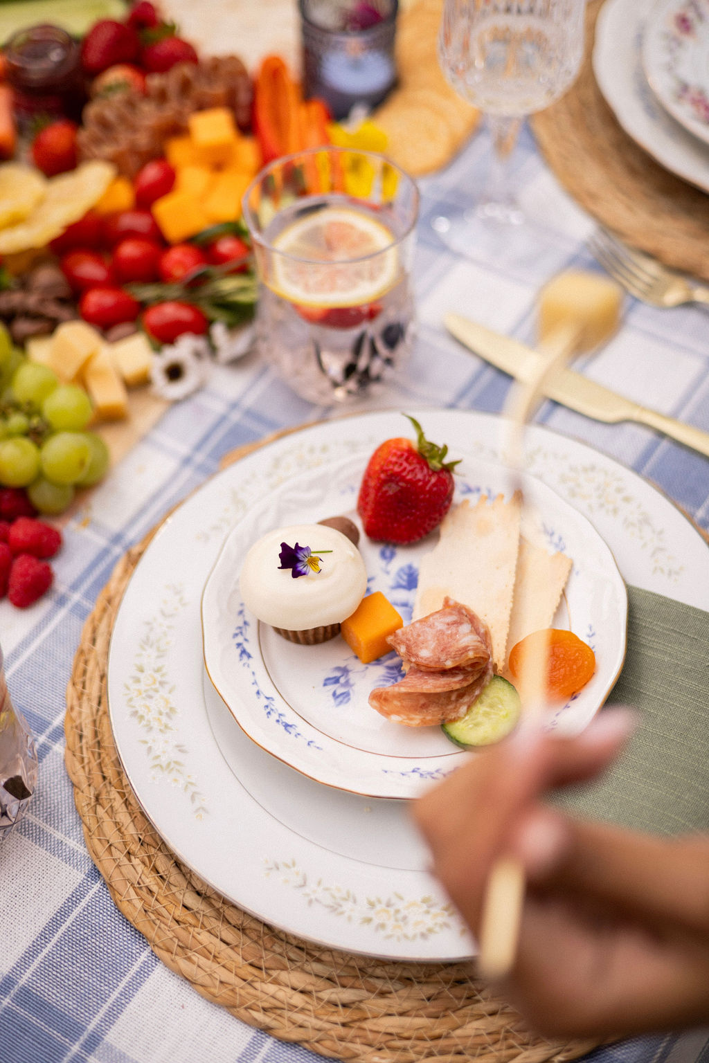 A vintage china place setting with a frosted cupcake, strawberry, salami and crackers