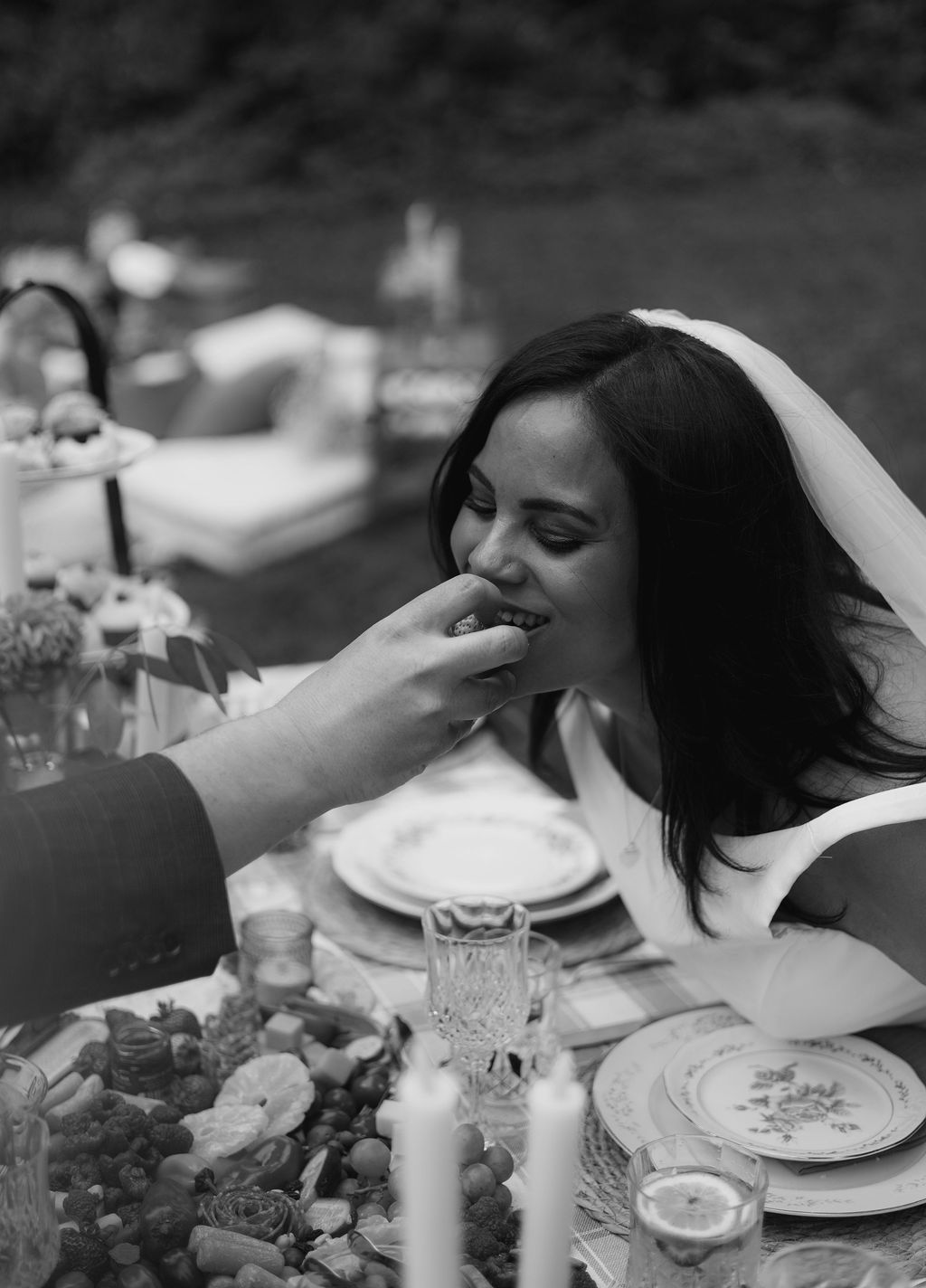 Black and white photograph of a groom feeding his bride at the picnic table at Cathedral Park
