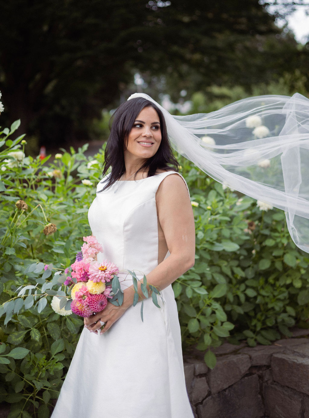 Bride holding a pink and yellow dahlia bouquet with her veil lifting in the wind at Cathedral Park