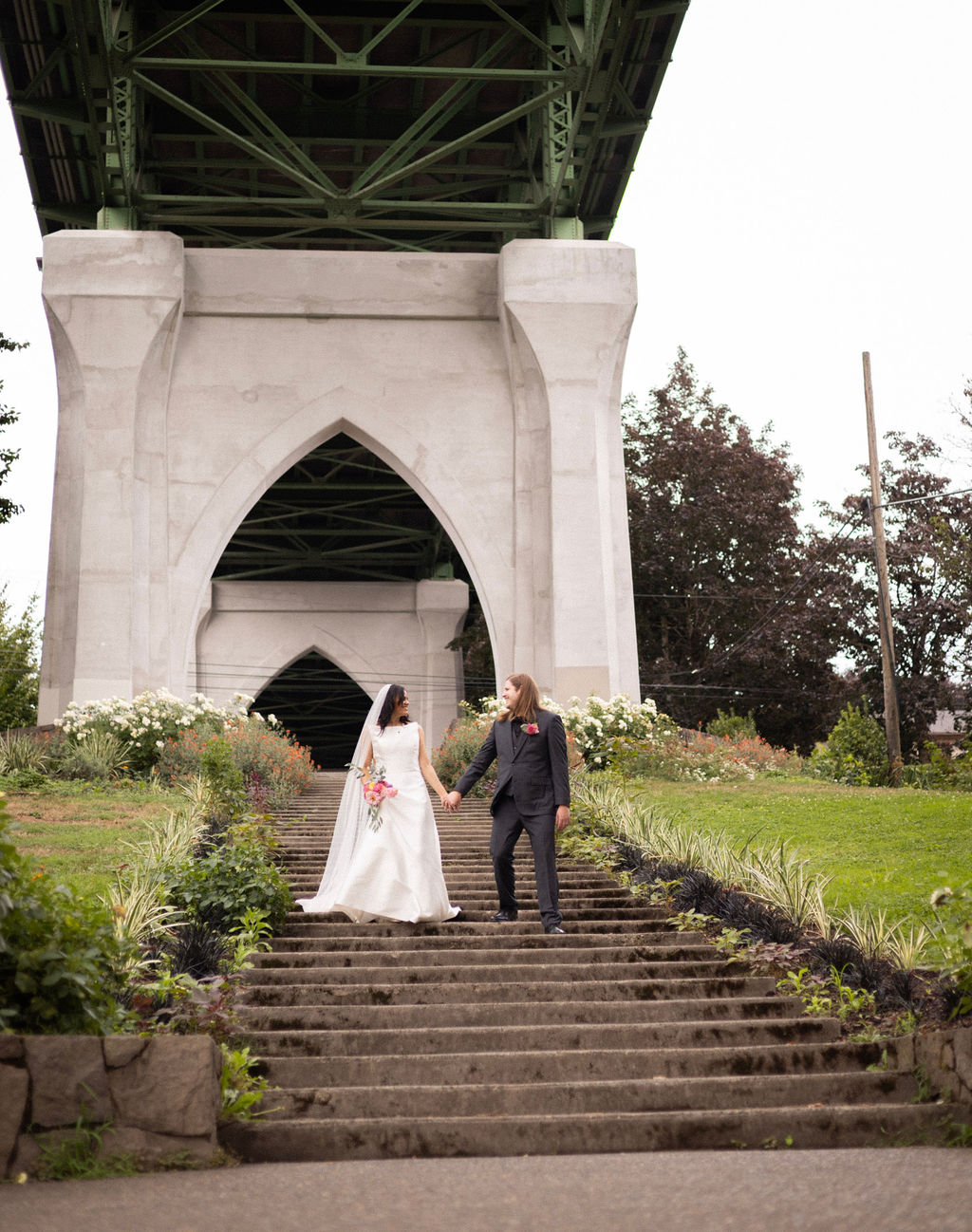 Newlyweds holding hands on the Cathedral Park staircase beneath the gothic arch of the St. Johns Bridge