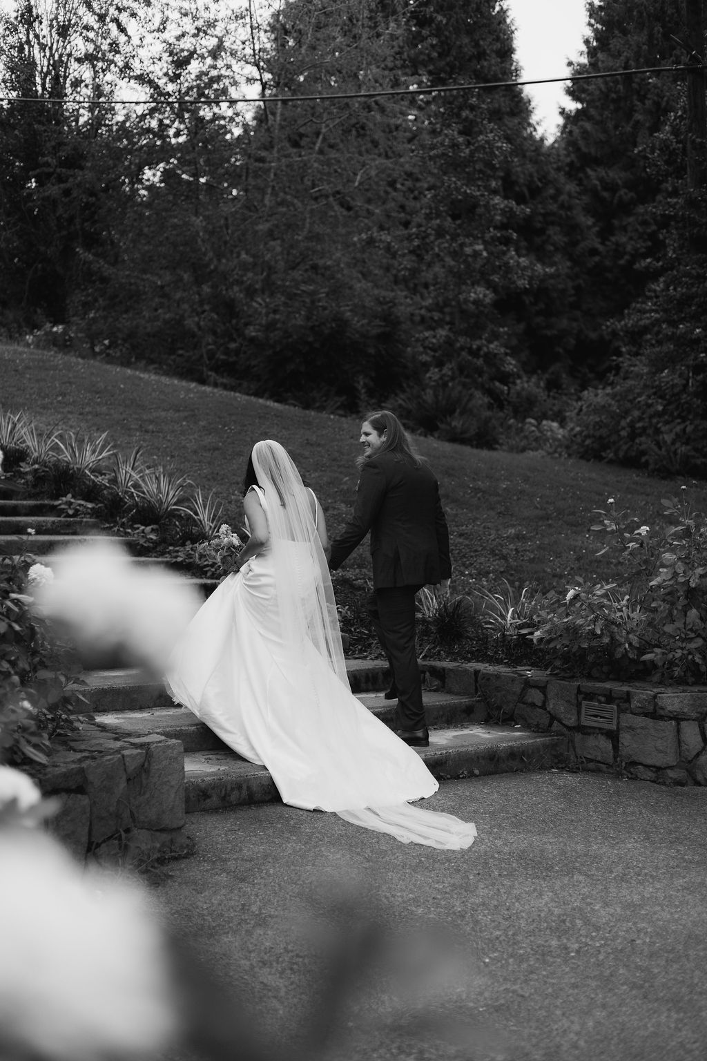 Black and white photograph of a bride and groom walking up the park steps, her veil trailing behind
